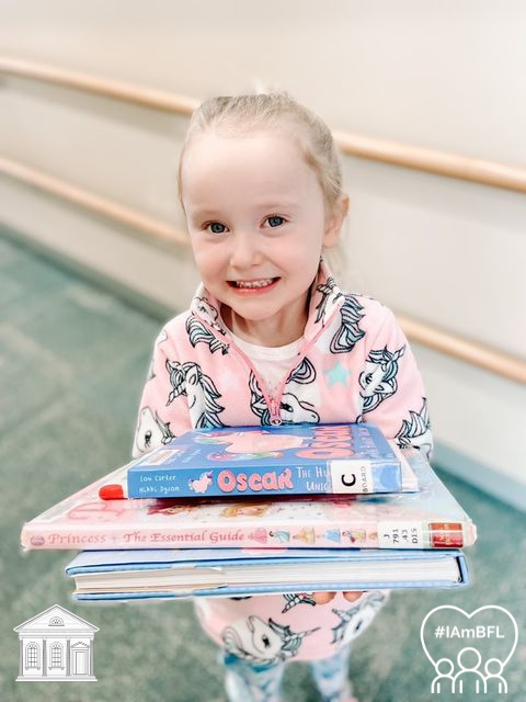 Caela smiling with a stack of books: Oscar Board Book, Princess: The Essential Guide, and a book that is unknown.