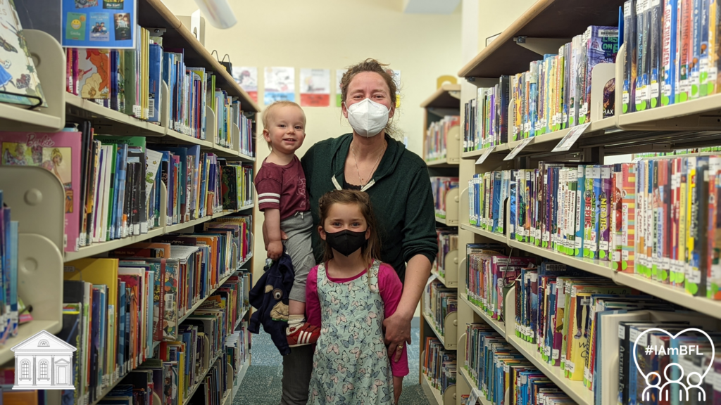 Elizabeth holding son Alexander and Charlotte standing in front of her. All are smiling, standing between the children's shelves.